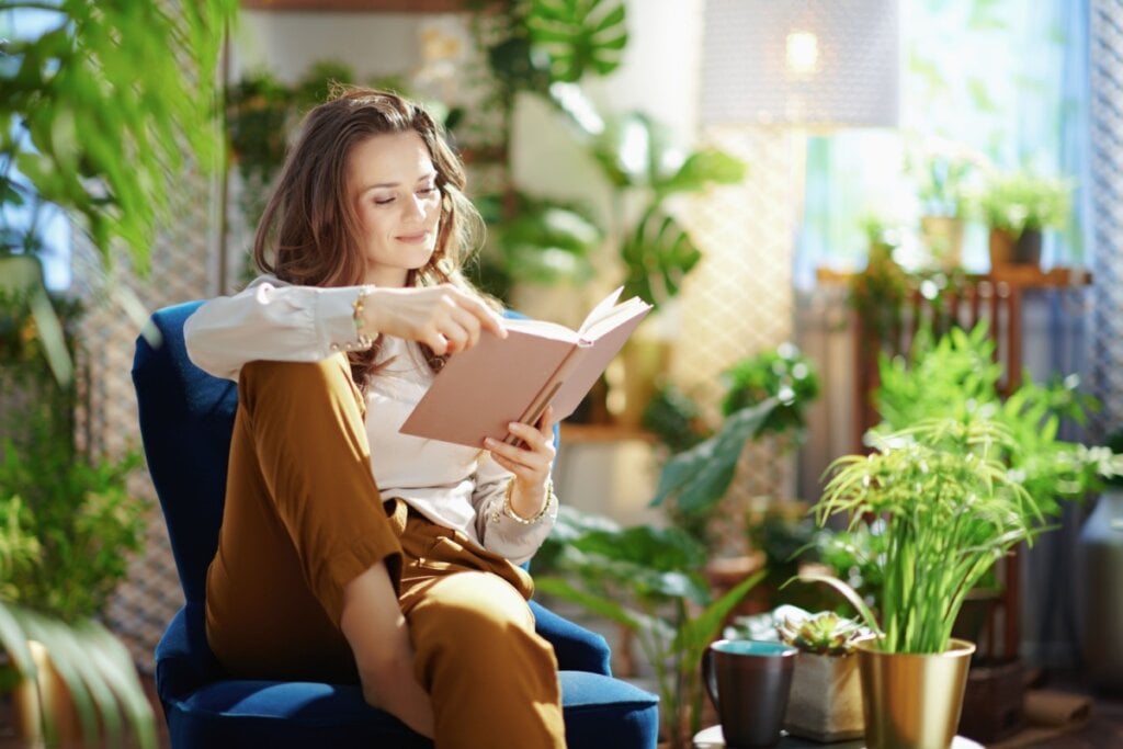 Mulher lendo livro sentada em uma poltrona azul em meio a diversas plantas com um semblante feliz