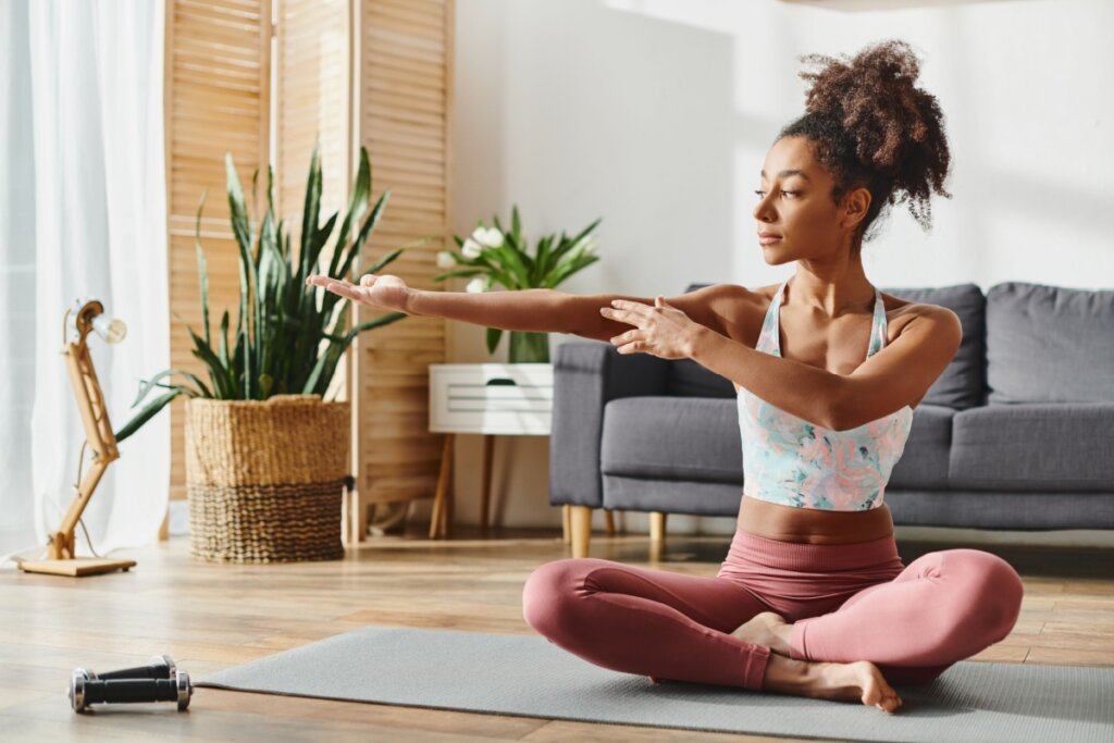 Mulher sentada em tapete na sala da casa praticando yoga. Ela está usando roupas próprias para exercício.