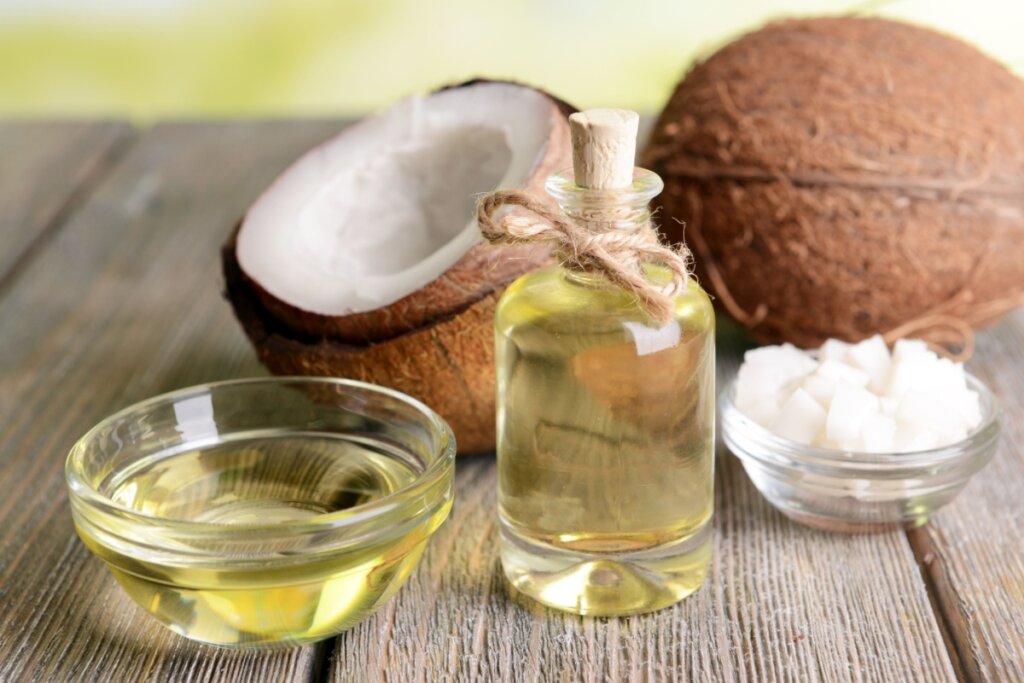 Em cima de uma mesa de madeira, estão uma cumbuca de vidro e um frasco com óleo de coco, ao lado um pote com coco ralado e atrás um coco partido ao meio e um coco inteiro