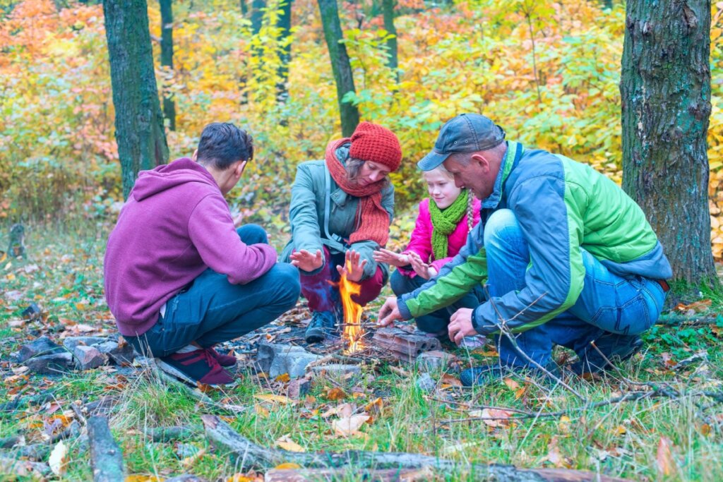 Uma família se aquecendo perto de uma fogueira; eles estão na floresta e vestindo roupas de frio.