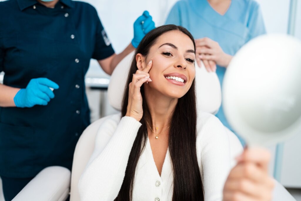 Mulher sorridente em uma clínica estética, segurando um espelho e apontando para o rosto, enquanto duas profissionais com luvas e uniformes médicos estão atrás