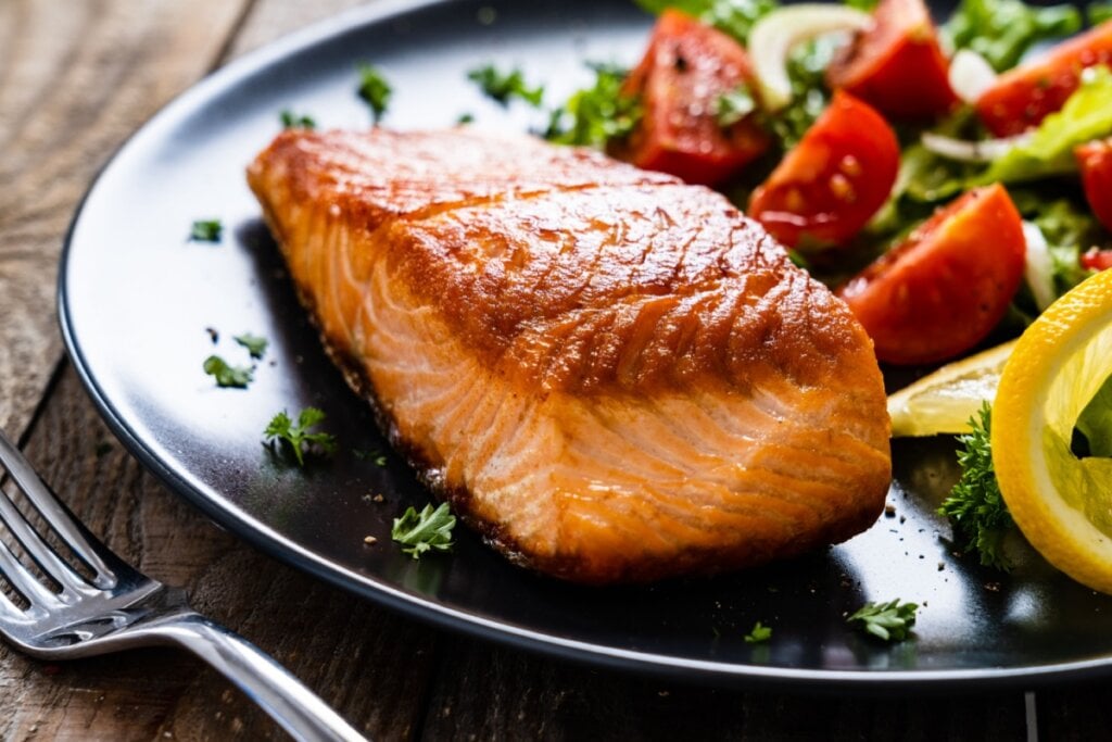 Salmão em prato preto com salada de tomate e folhas verdes