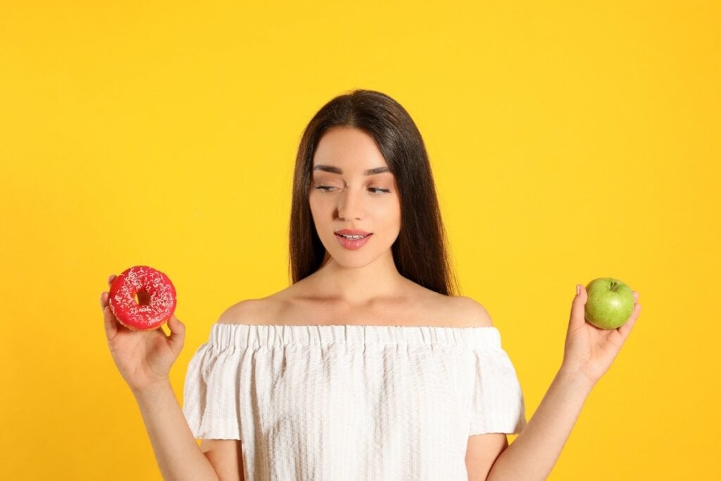 Mulher usando blusa branca e segurando um donut e uma maçã em cada uma das mãos