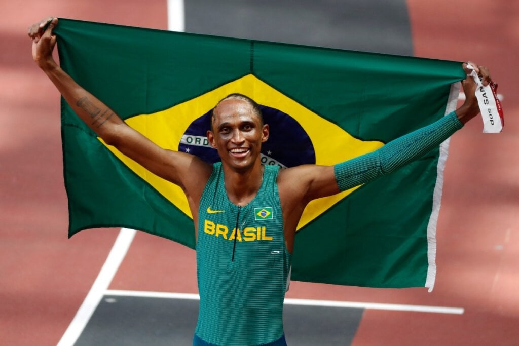 Alison dos Santos sorrindo segurando a bandeira no Brasil nas costas 