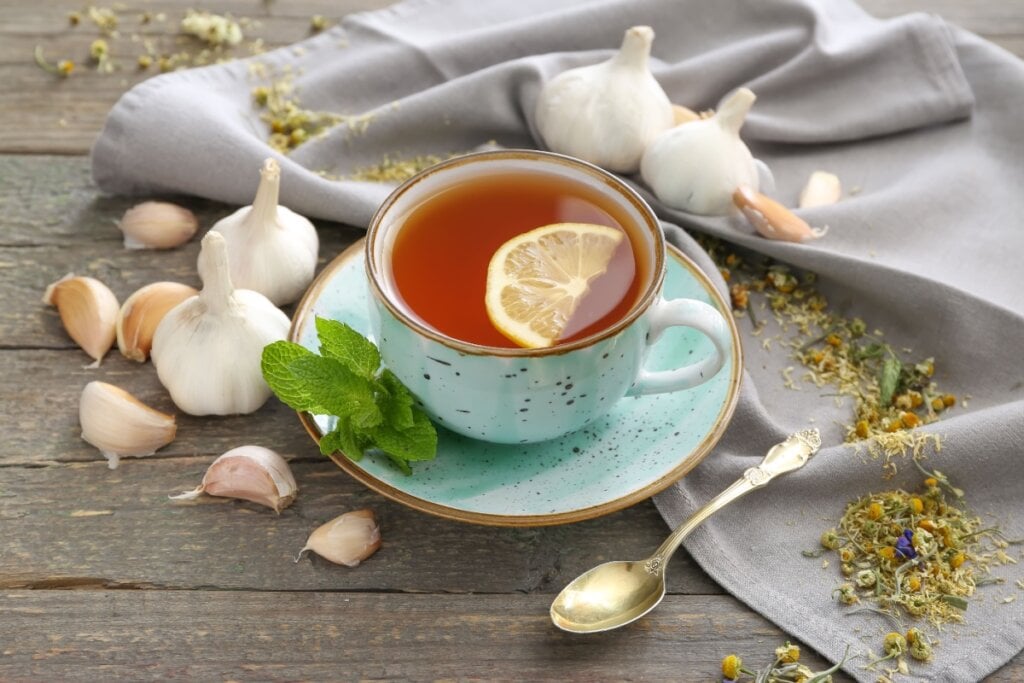 Xícara de chá de hortelã com limão, cercada por cabeças de alho, folhas de hortelã e um pano cinza
