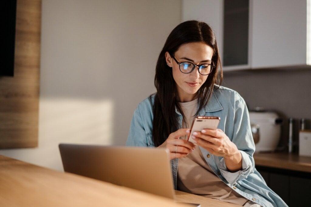 Mulher de cabelo liso usando óculos sentada em frente a um notebook mexendo no celular 