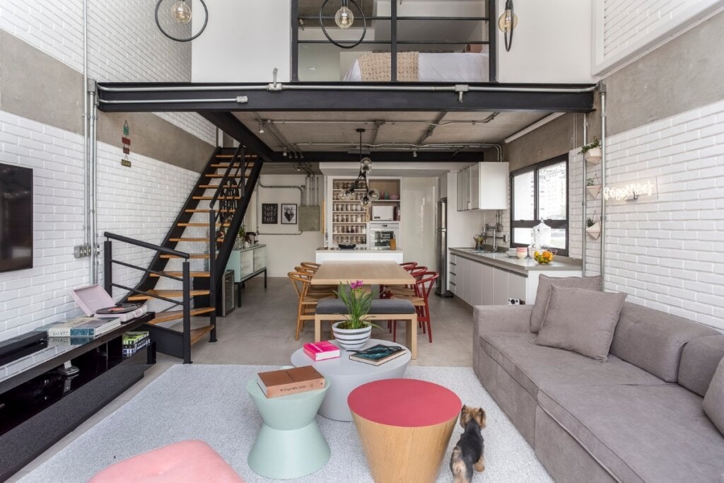 Loft com escada de metal, paredes brancas, sofá cinza e mesinhas e bancos em formato geométrico 