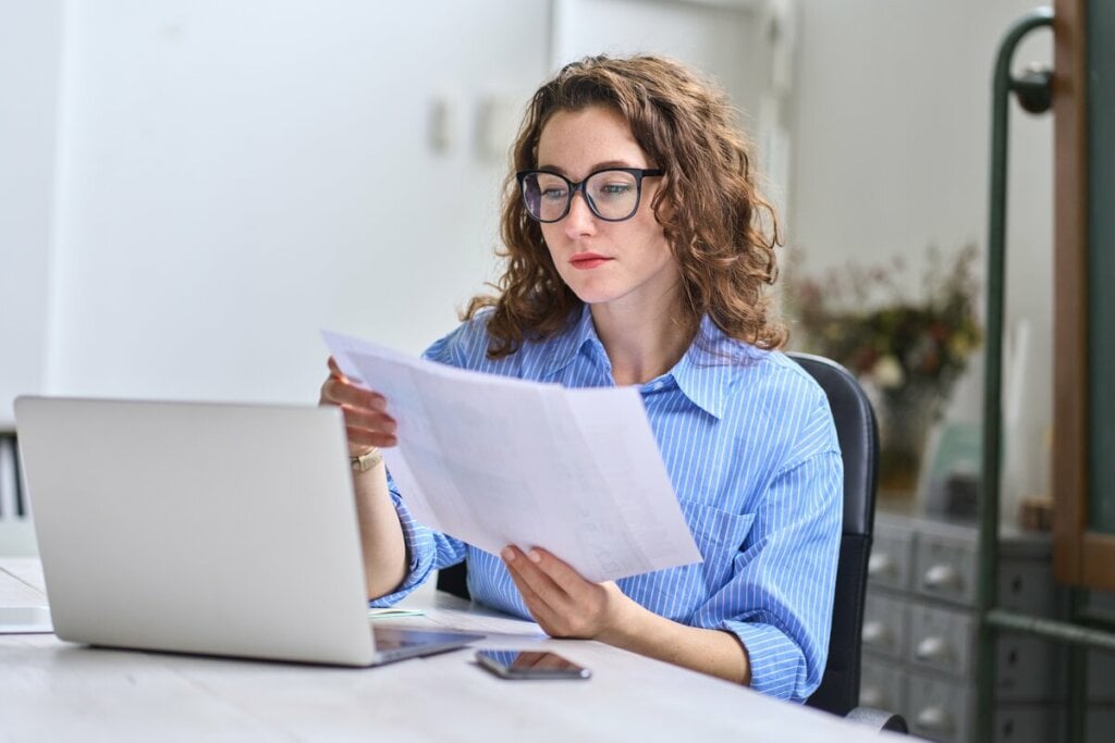 Mulher usando uma camisa azul sentada em frente a um notebook olhando um papel