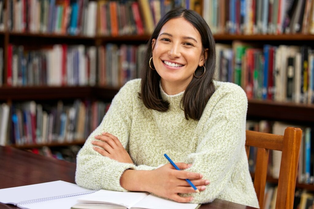 Mulher sentada em uma biblioteca sorrindo com os braços cruzados em cima de uma mesa com livros e papéis