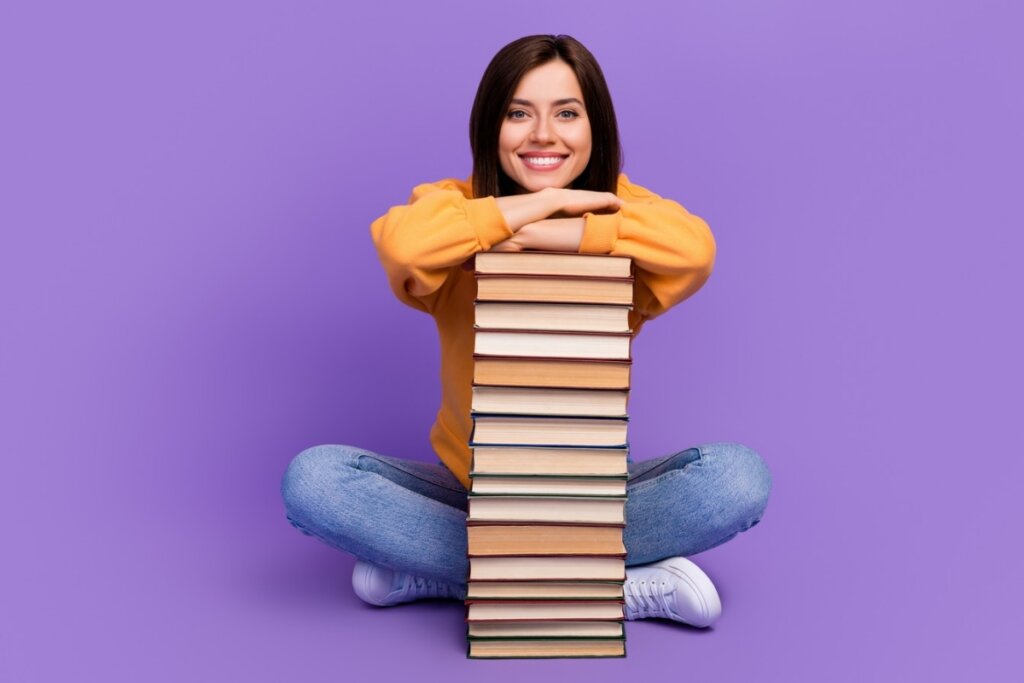 Mulher sentada sorrindo apoiando os dois braços em uma pilha de livros em um fundo roxo