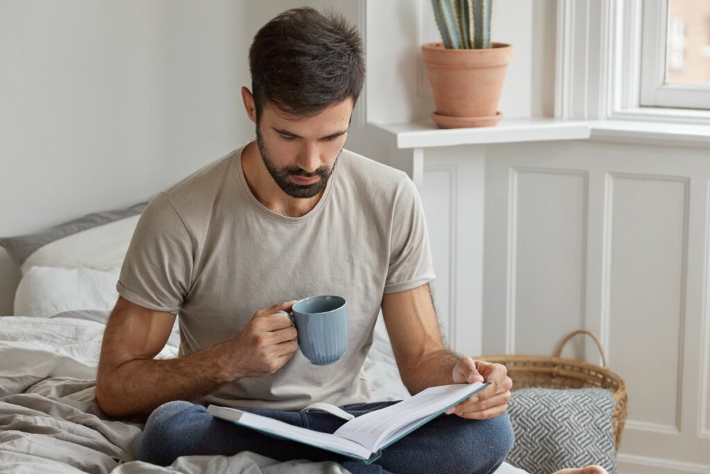 Homem sentado em uma cama segurando uma caneca e lendo um livro