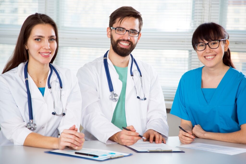 Dois médicos vestindo jaleco e uma enfermeira vestindo uniforme azul sentados com os braços cruzados em uma mesa