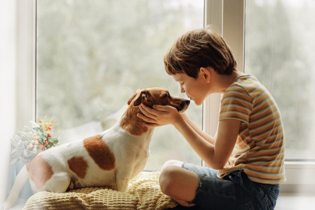 Menino dando beijo no rosto de um cachorro em frente a uma janela