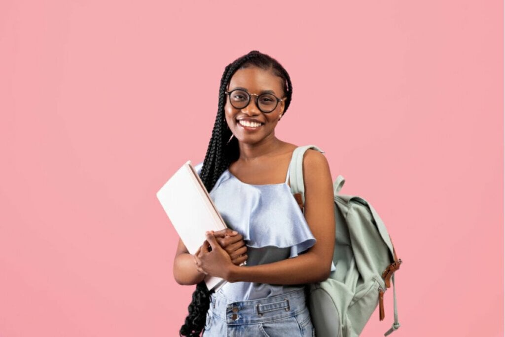 Mulher segurando livros e com mochila nas costas em frente a um fundo rosa