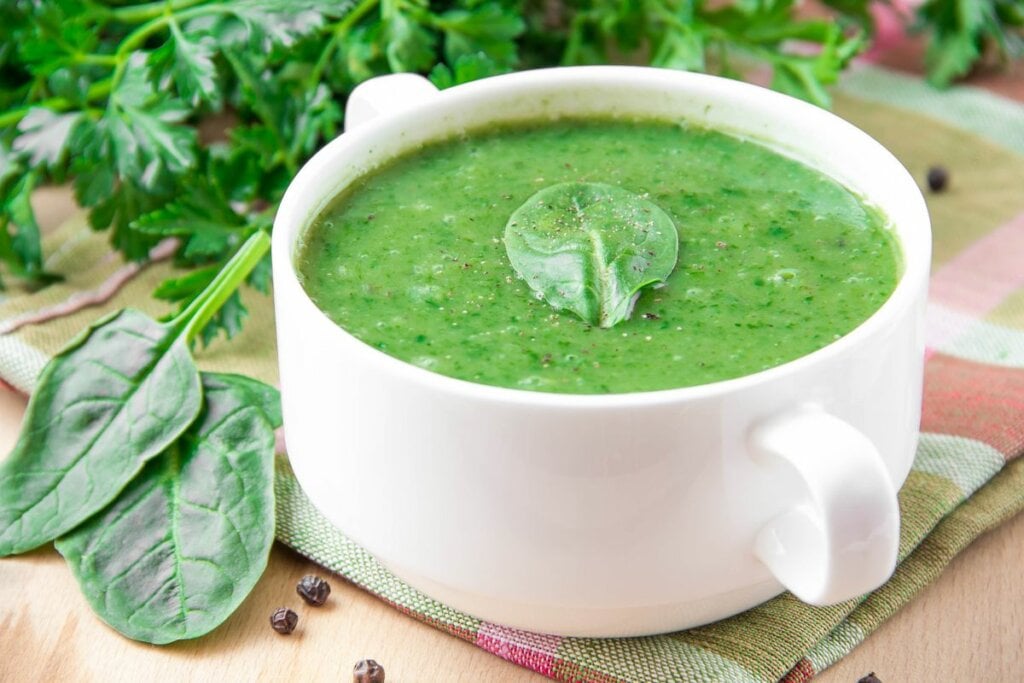 Panela de porcelana branca com sopa verde em cima de uma mesa de madeira com folhas verdes