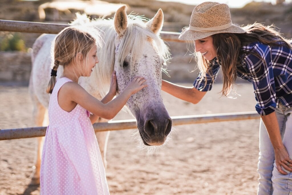 Menina fazendo carinho em um cavalo branco com uma moça do lado 