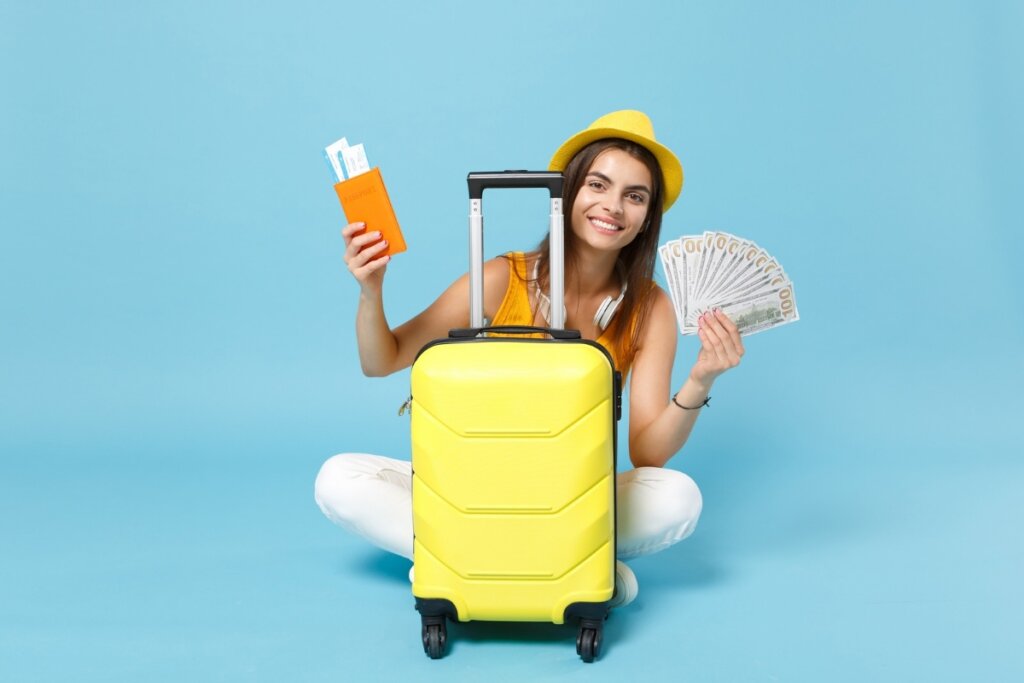 Mulher usando chapéu sentada com uma mala amarela na sua frente feliz segurando um passaporte e dinheiro