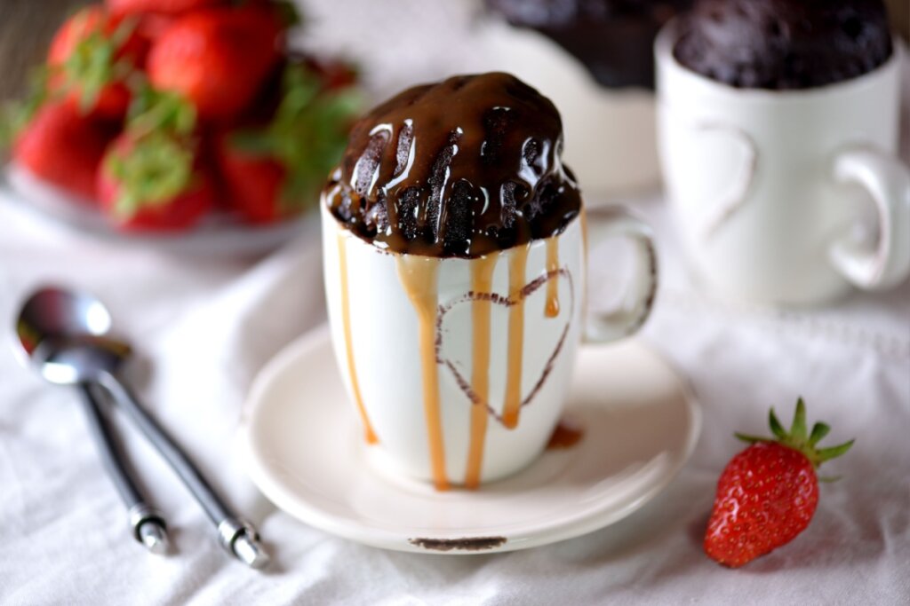 Bolo de chocolate em caneca branca com coração e calda em cima. Há morangos ao fundo