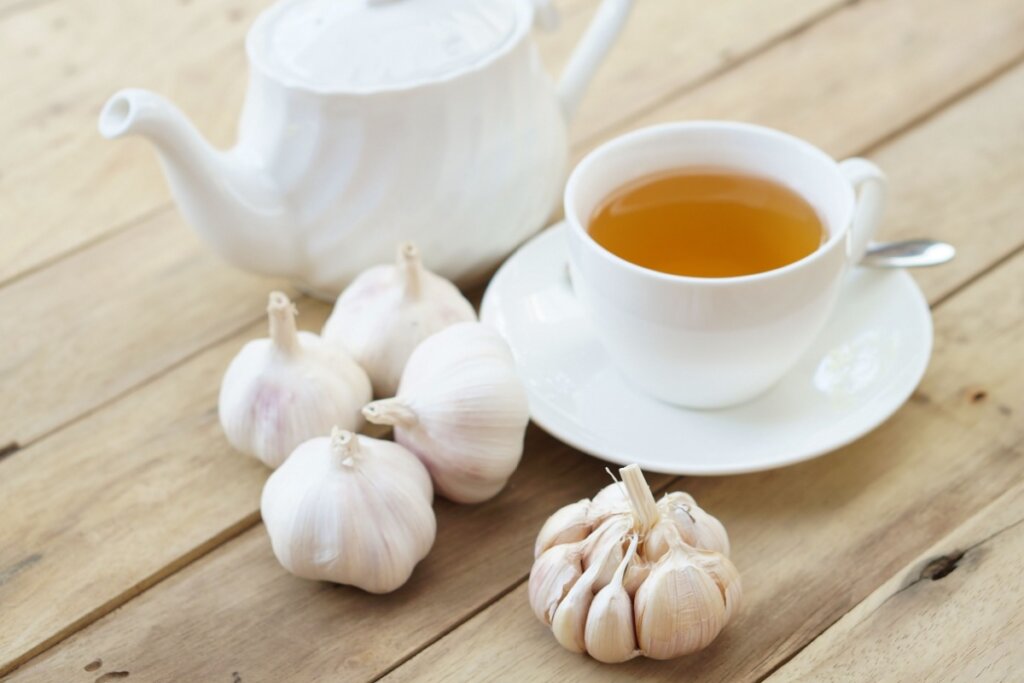 Bule e xícara de porcelana branca com chá de alho, e pedaços de alho ao redor em uma mesa de madeira