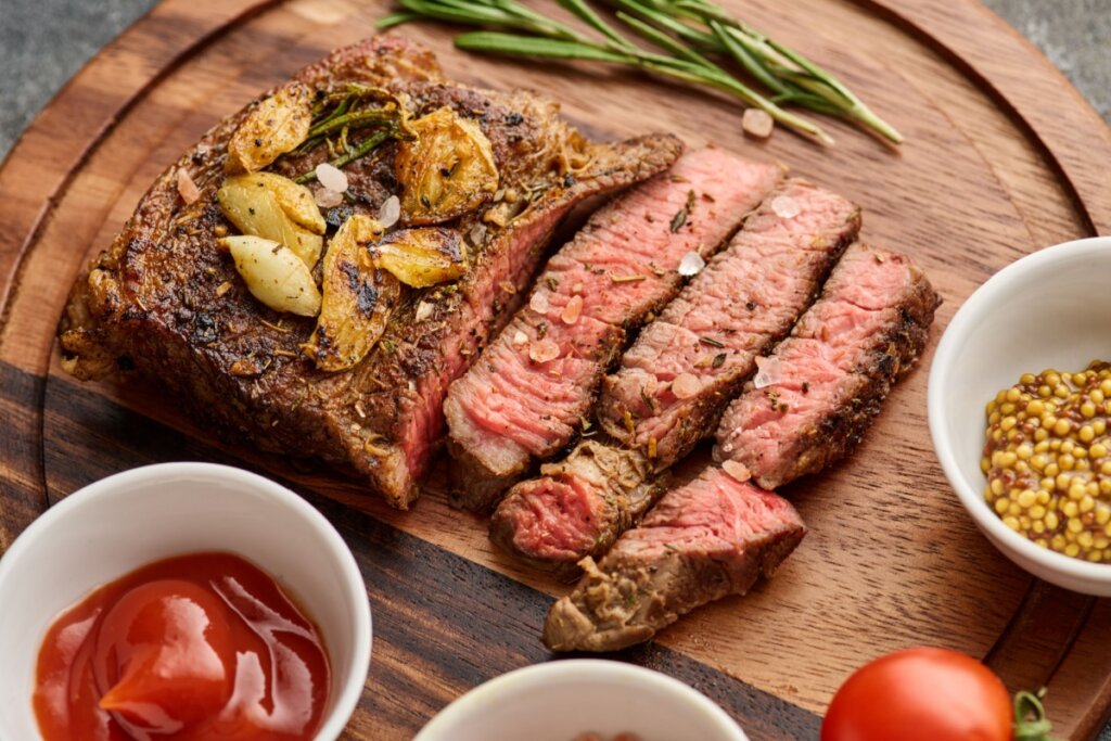 Bife cortado em uma tábua de madeira, com cumbuca com molhos e temperos ao redor. Em cima do bife há pedaços de alho.