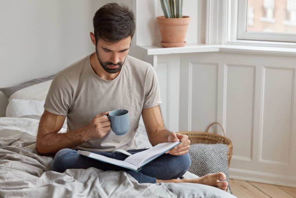 homem lendo livro segurando caneca sentando em uma cama