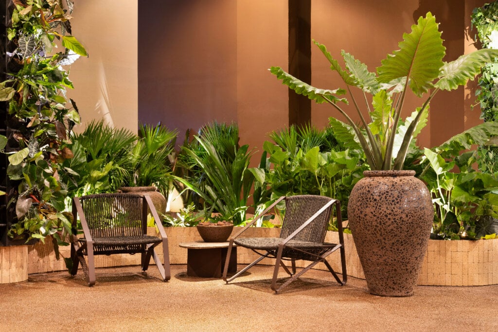 Jardim de inverno com diversos vasos de planta ao fundo e com duas cadeiras e uma mesa de apoio na frente e do lado um vaso grande de plantas 