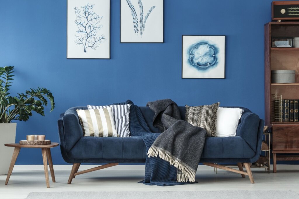 Sala de estar com sofá azul, cobertores e almofadas listradas e estampadas, decorada com quadros de arte em uma parede azul, acompanhada de uma mesa lateral de madeira com velas e uma estante com livros