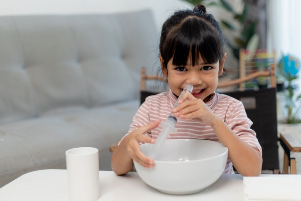 Menina sorrindo com seringa com soro perto do nariz e pote branco em mesa