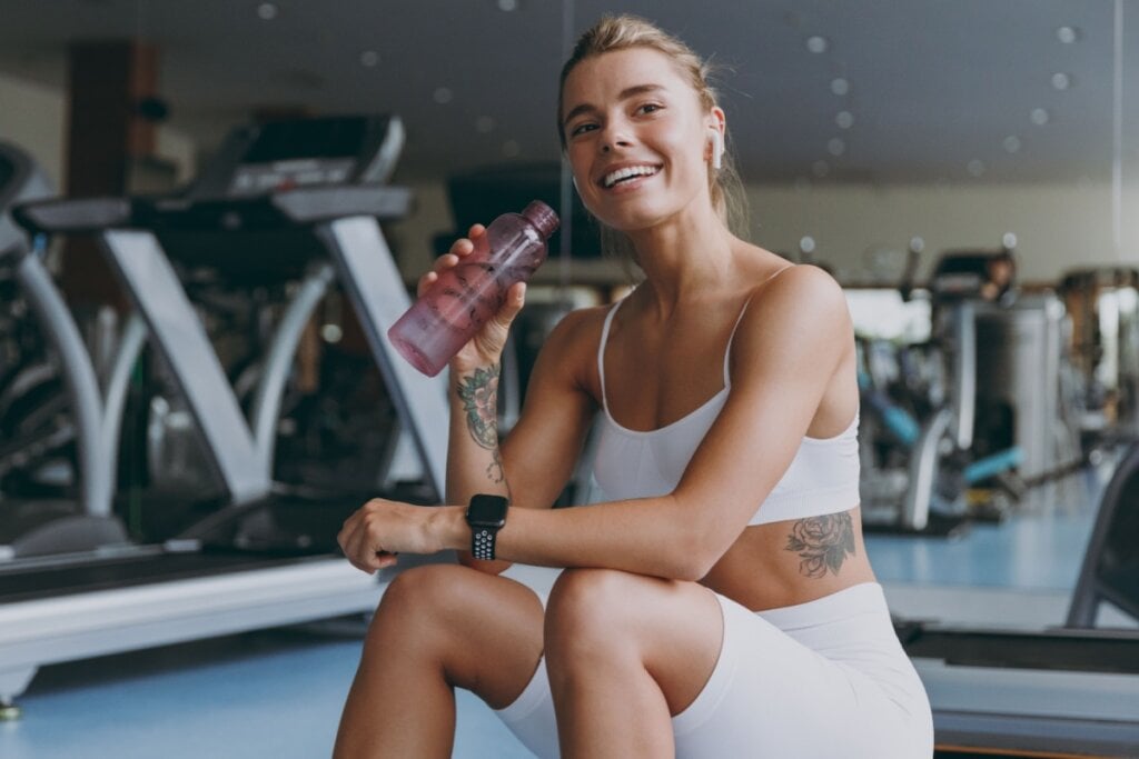 Mulher em academia sorrindo enquanto bebe água de garrafinha 