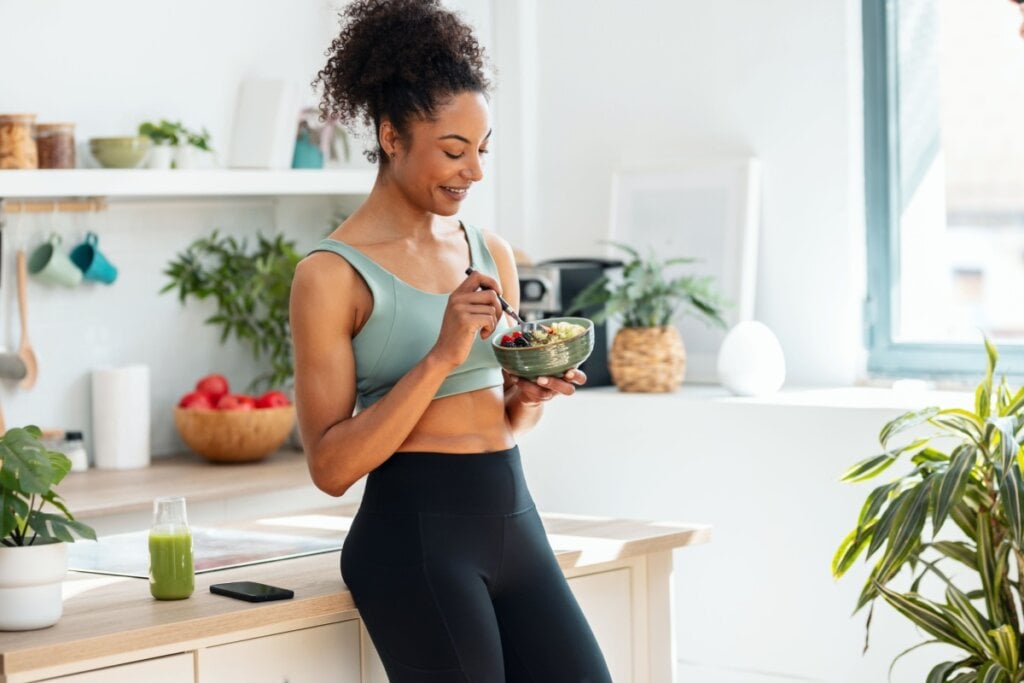Mulher apoiada em bancada em cozinha comendo comida saudável em um bowl