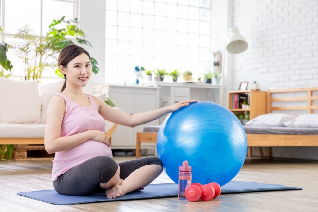 Mulher grávida sentada no chão sobre tapete de yoga segurando bola de pilates azul ao lado