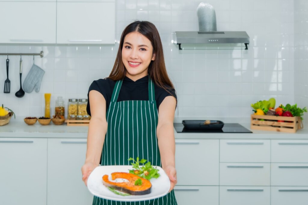 Mulher na cozinha com um prato com peixe nas mãos.