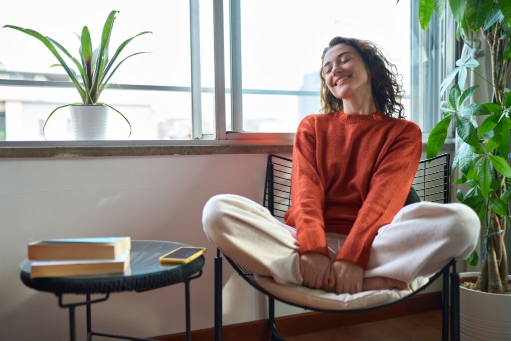 Mulher sentada em uma cadeira com um semblante feliz, ao lado está uma mesa com livros e um celular e, no parapeito da janela, há um vaso de plantas