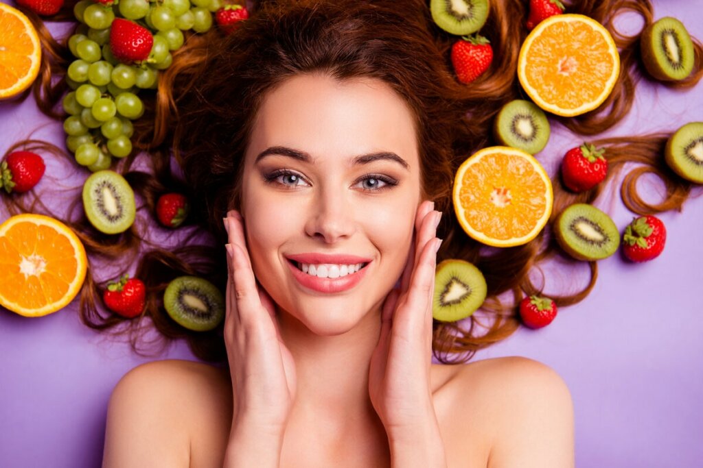 Mulher sorrindo deitada sobre um fundo roxo com as mãos no rosto e frutas ao redor