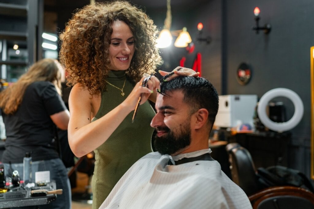 Mulher em pé com um vestido verde cortando o cabelo de um homem sentado