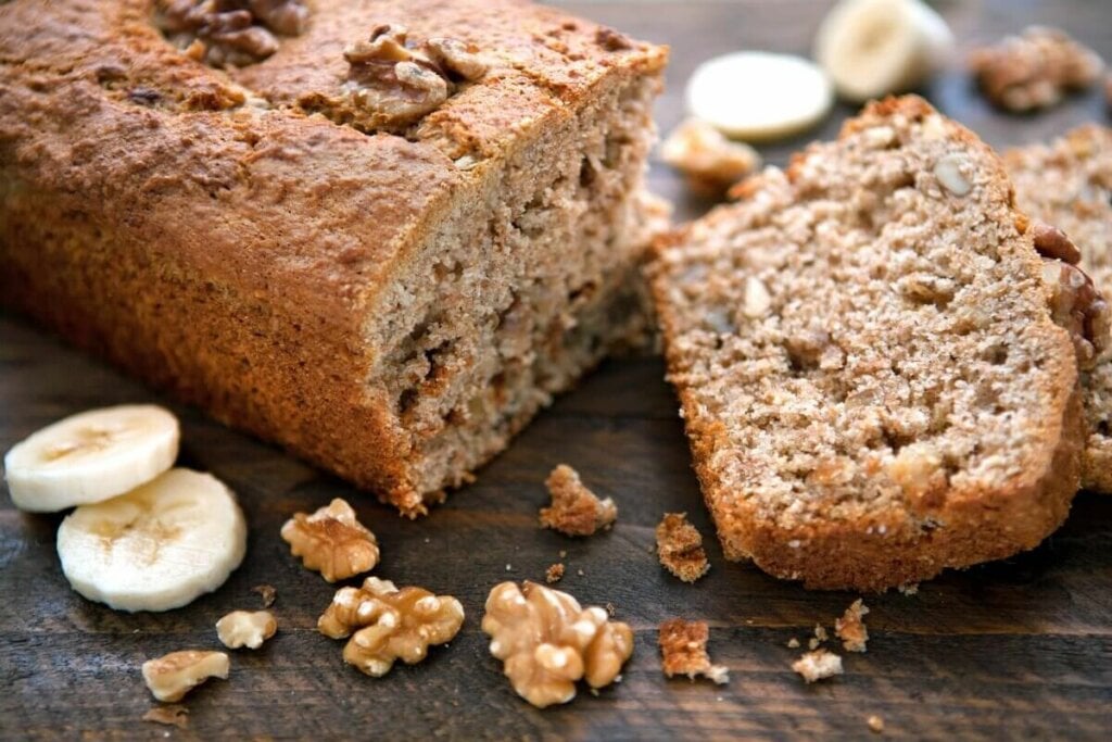 Bolo de banana com nozes em cima de uma mesa de madeira com rodelas de banana e pedaços de nozes