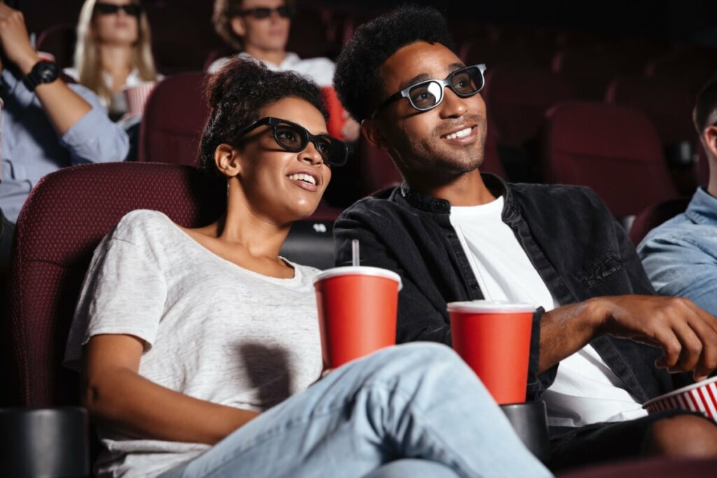 Casal sorrindo no cinema comendo pipoca e bebendo em copo vermelho