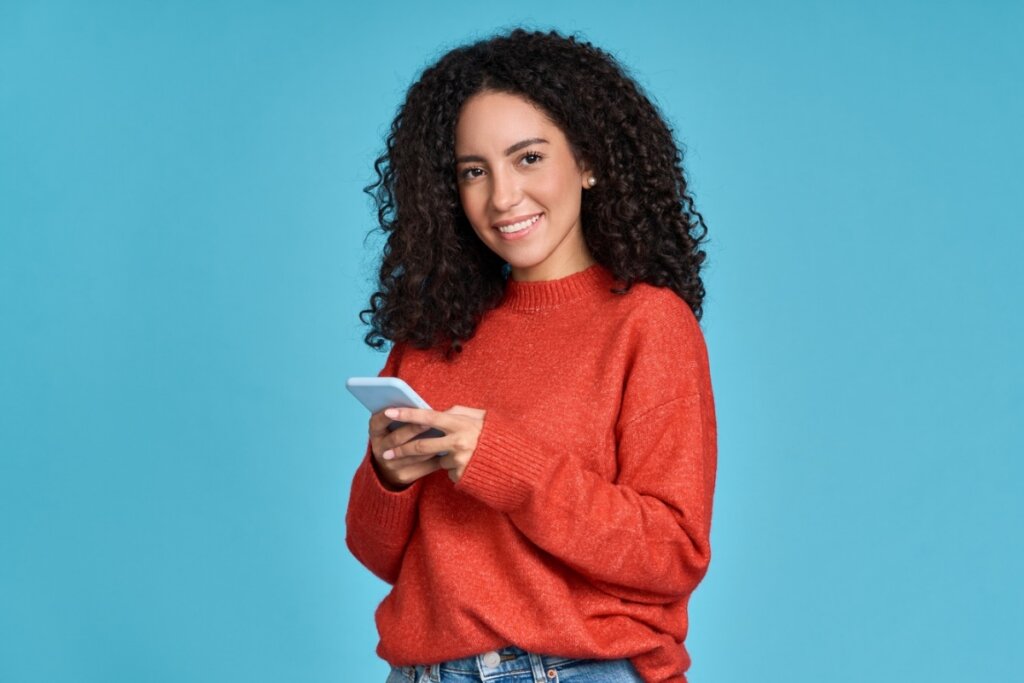 Mulher usando blusa vermelha de manga comprida em um fundo azul, sorrindo e mexendo em um celular