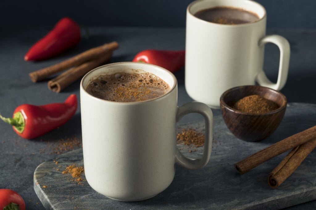 Duas canecas brancas de chocolate quente cremoso com pimenta e especiarias, ao lado de pimentas vermelhas frescas, paus de canela e um recipiente de madeira com especiarias