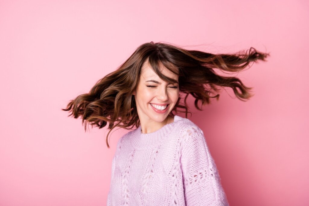 Mulher vestindo blusa de tricô rosa, sorrindo em frente a um fundo rosa com os cabelos voando