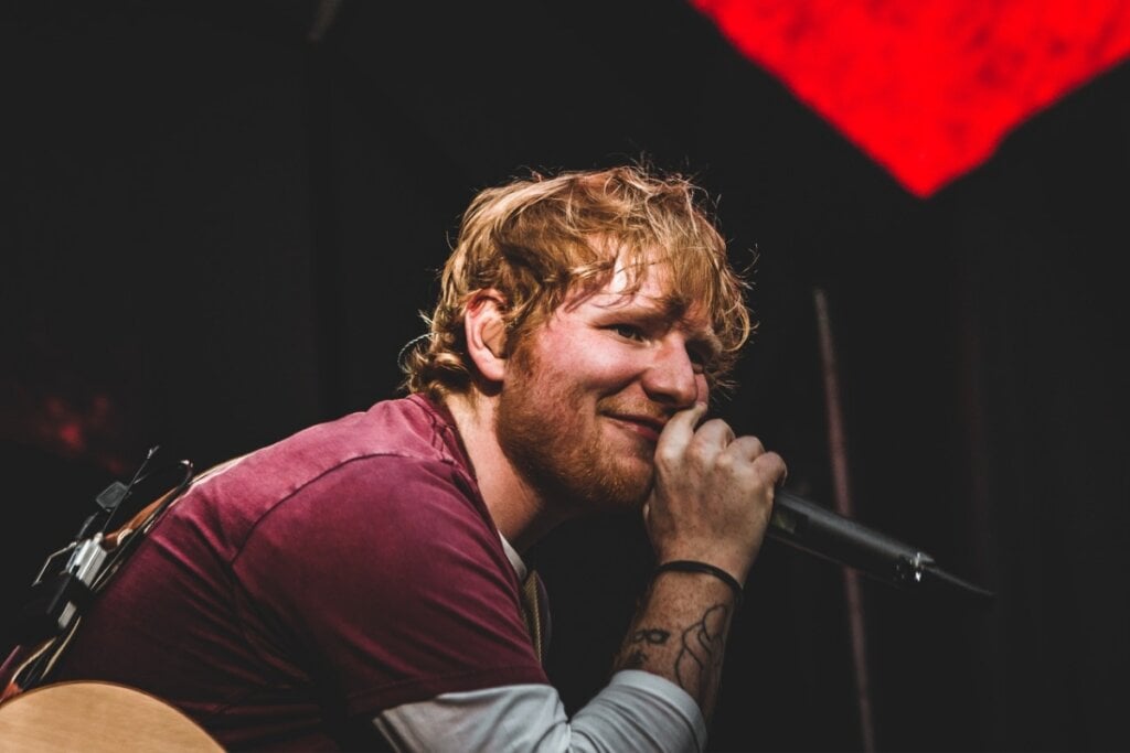 Ed Sheeran vestindo uma camisa vermelha e segurando um microfone no palco 