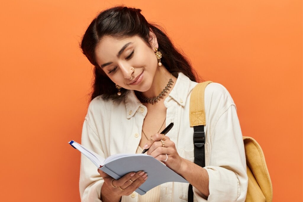 Mulher com mochila amarela nas costas segurando um livro e escrevendo nele