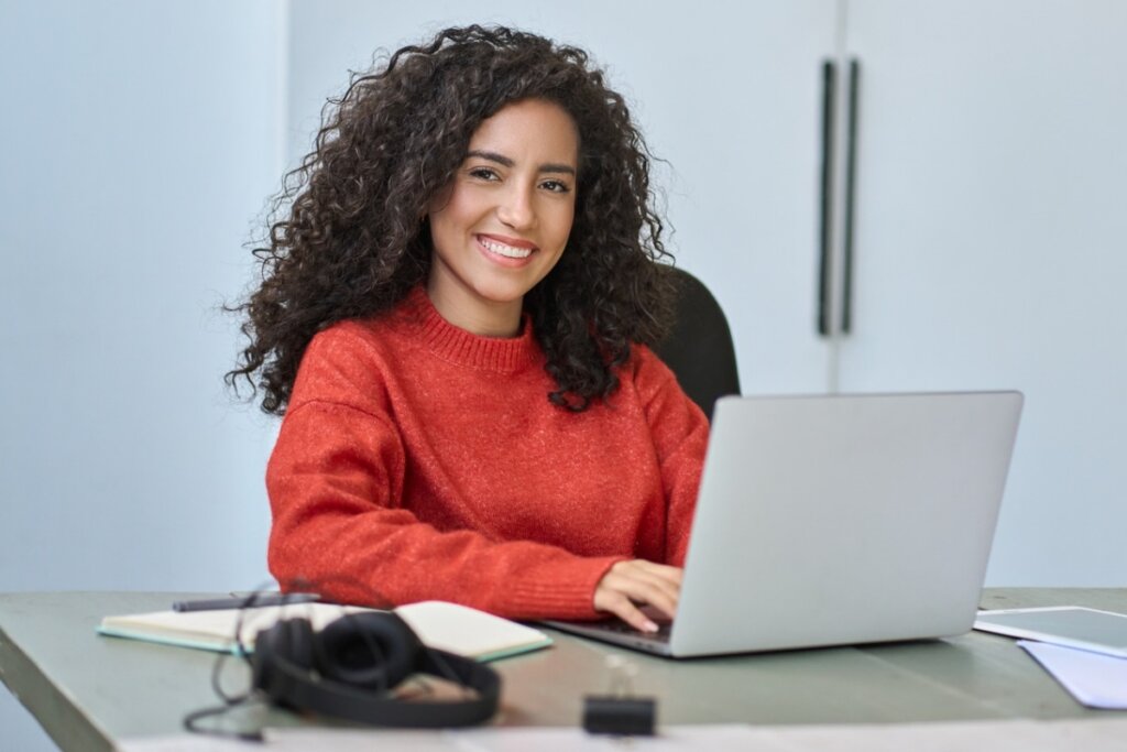 Mulher de cabelo cacheado sentada sorrindo e mexendo em um notebook