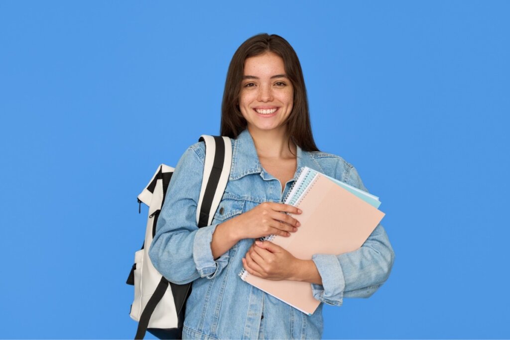 Menina com uma mochila branca e preta nas costas, segurando livros e sorrindo em frente a um fundo azul-claro