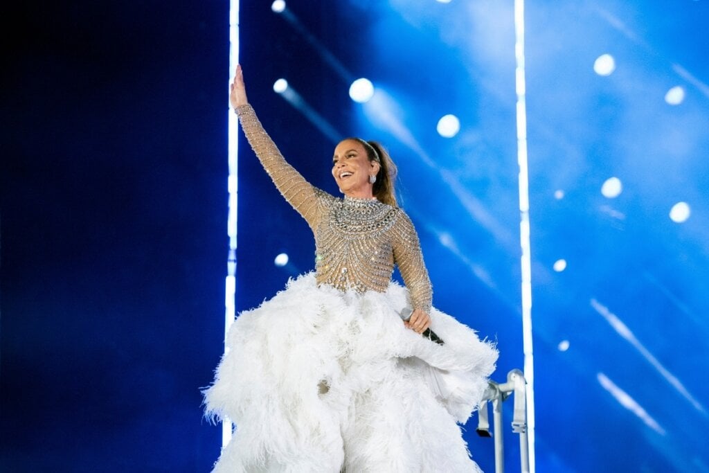 Ivete Sangalo usando um vestido com brilho na parte de cima e plumas branca na saia em um palco com luzes azuis 