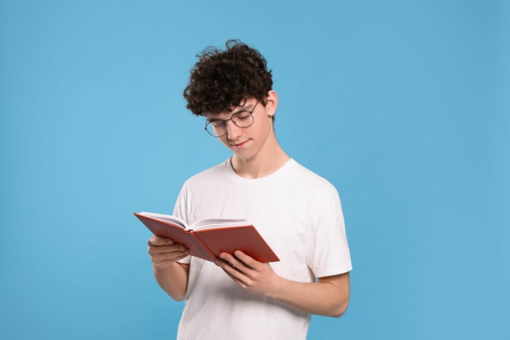 Menino usando uma blusa branca e óculos de grau lendo um livro em frente a um fundo azul