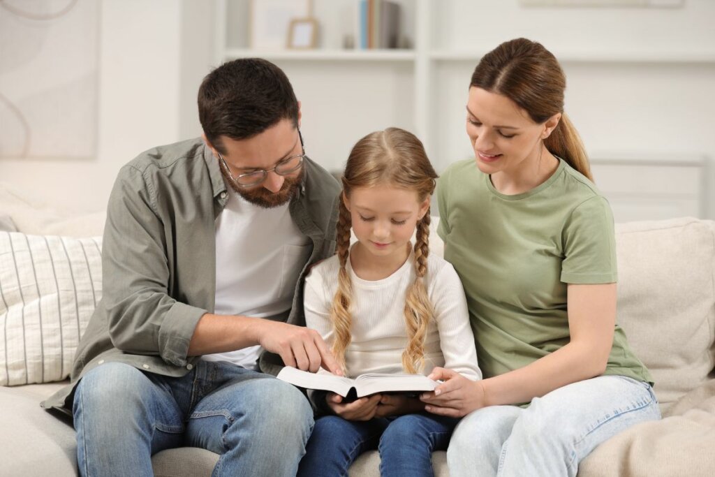 Homem, mulher e menina sentados em um sofá lendo um livro