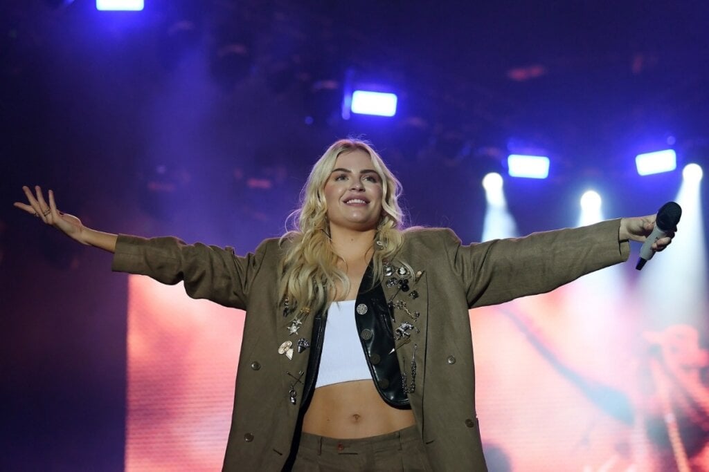 Luiza Sonza vestindo um cropped branco com camisa verde por cima em cima de um palco com os braços abertos 