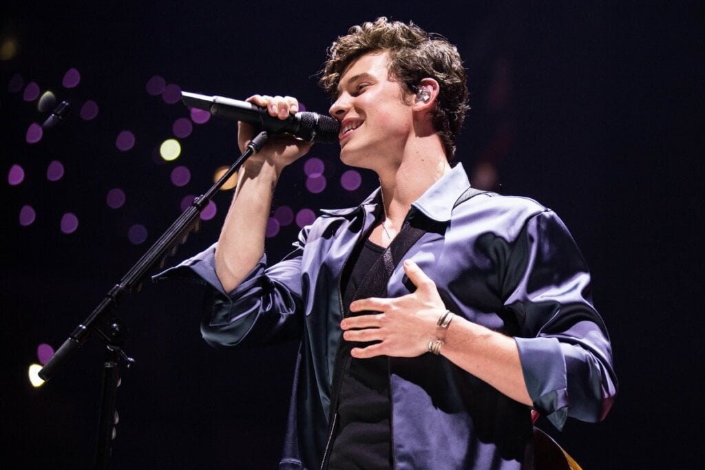 Shawn Mendes usando uma camisa azul-escuro em cima de uma palco segurando um microfone preto e cantando 