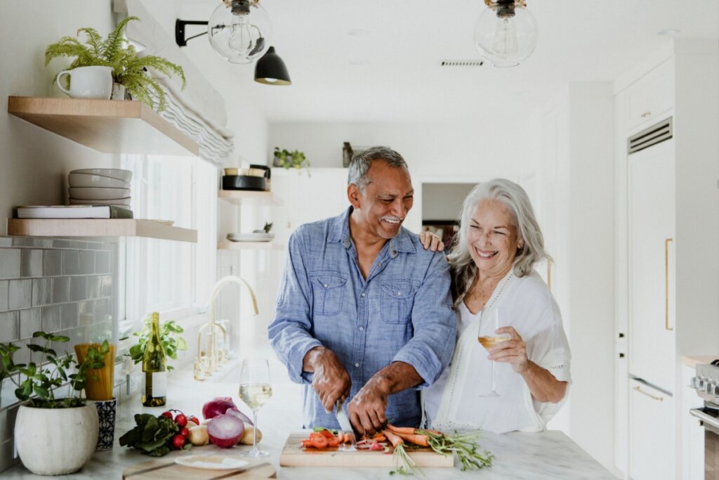 casal de idosos sorrindo na cozinha. homem está cortando cenoura em bancada